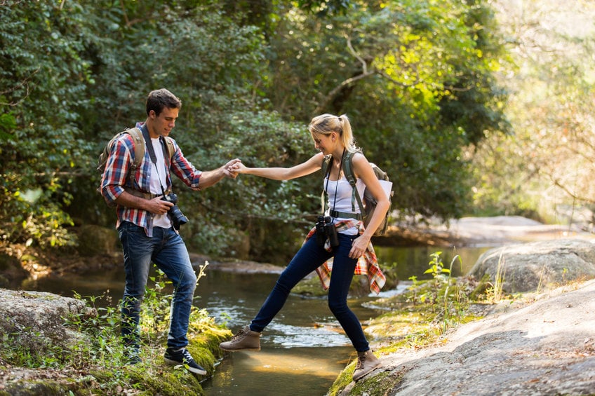 young man helping girlfriend crossing stream at mountain valley