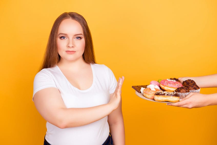 I'm against eating products containing fat! Will-powered woman wearing white tshirt is refusing to consume tasty delicious sweets on a plate, isolated on bright yellow background