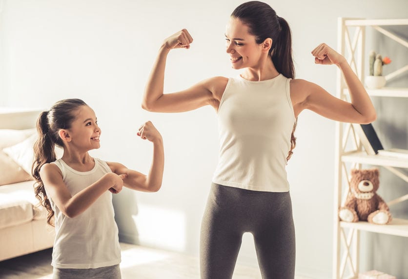 Beautiful young woman and charming little daughter are showing their biceps and smiling while working out at home