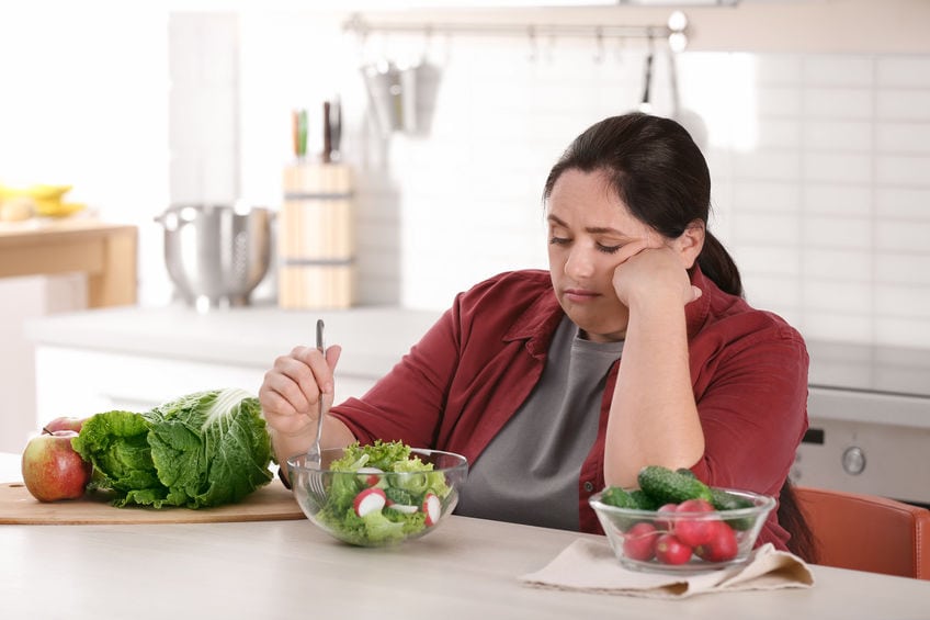 Unhappy woman eating vegetable salad at table in kitchen. Healthy diet