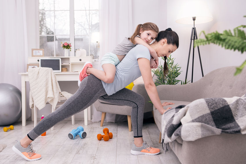 In high spirits. Determined athletic mother training at home and holding her daughter on her back