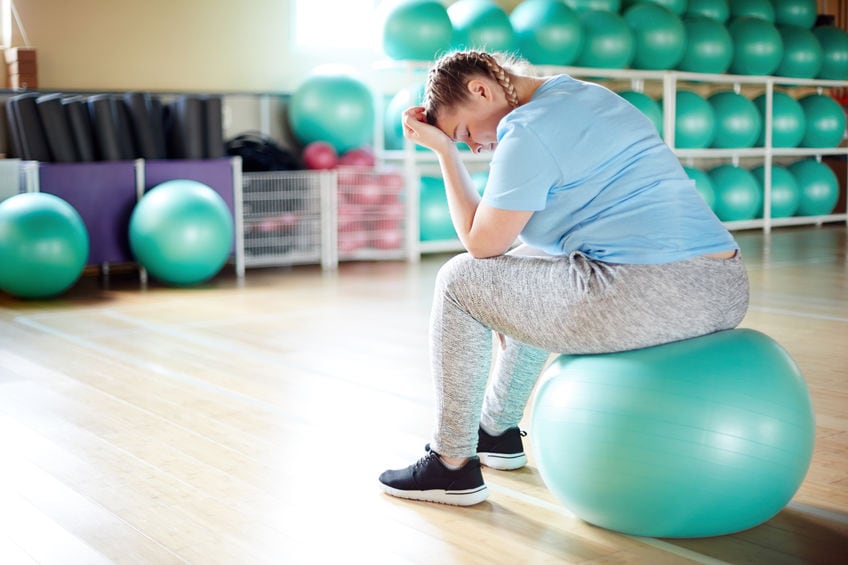 Young plus-sized woman sitting on fitball in gym and thinking about her bad fitness results